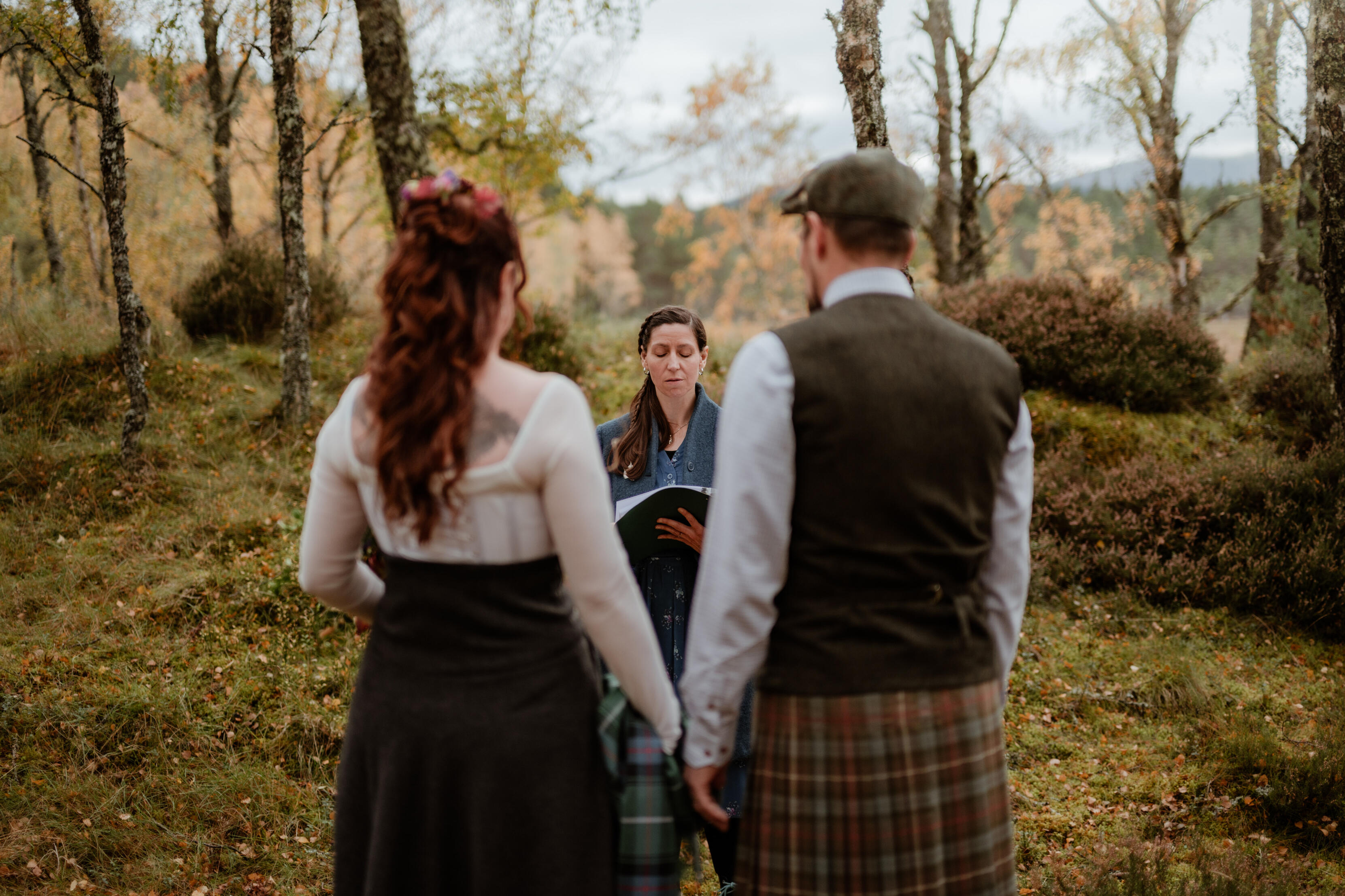 In Ceremony Emily stands in front of a couple who are facing her, with their backs to the photographer. Emily's eyes are closed and she holds the script in front of her. It is autumn and behind her is a small hill covered in moss and blaeberries, with heather and birc
